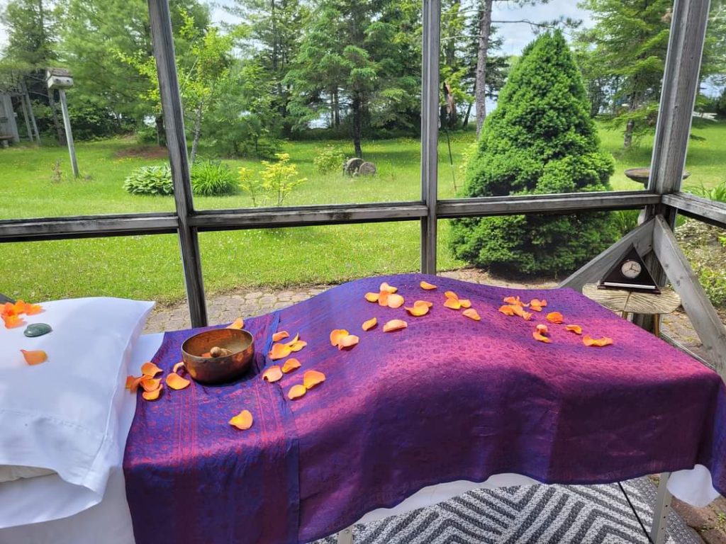 Healing space - Healing table covered with inviting woven fabric, rose petals and a sound healing bowl, in a window filled room.